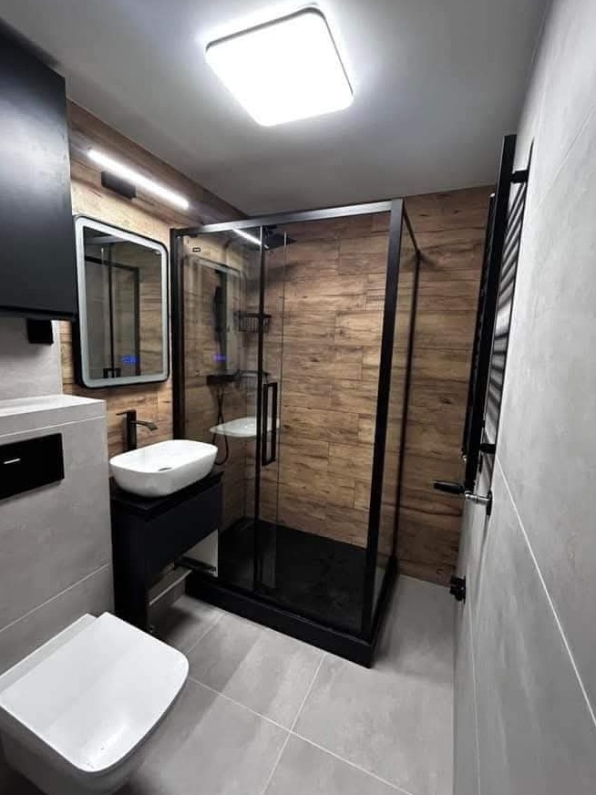 Nowoczesna łazienka z kabiną prysznicową typu walk-in z czarnymi profilami, umywalką nablatową na czarnej szafce, lustrem z podświetleniem LED i płytkami imitującymi drewno na ścianie kabiny.
