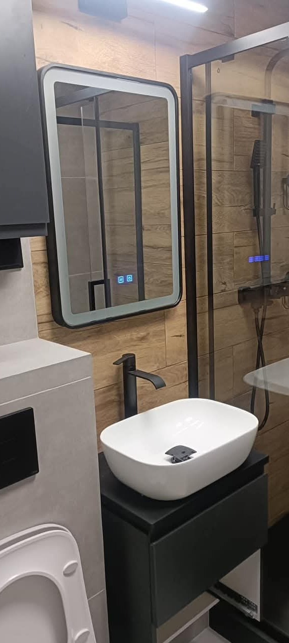 Nowoczesna łazienka z umywalką nablatową, czarną armaturą i podświetlanym lustrem z wyświetlaczem, kabina prysznicowa z drewnopodobnymi płytkami na ścianie.