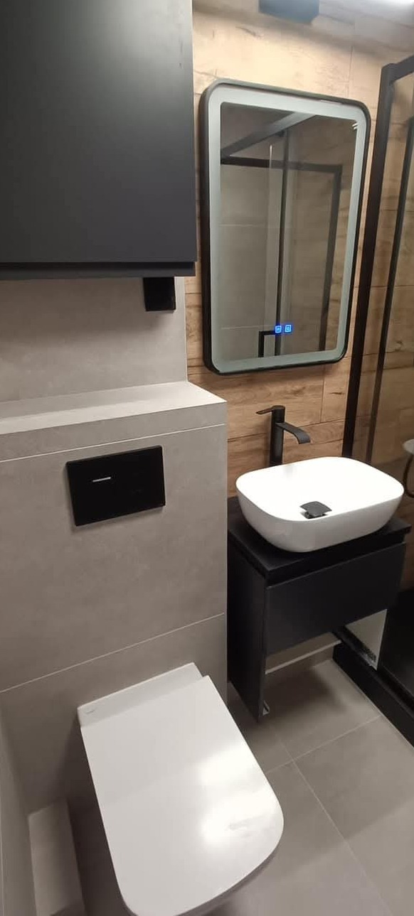 Nowoczesna łazienka z wiszącą toaletą, umywalką nablatową na czarnej szafce i podświetlanym lustrem w czarnej ramie, wykończona płytkami imitującymi drewno i szarym gresem.