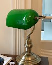 Klasyczna lampka biurkowa z zielonym, szklanym kloszem i mosiężną podstawą, ustawiona na drewnianym biurku, w tle okno z rozmytym widokiem.