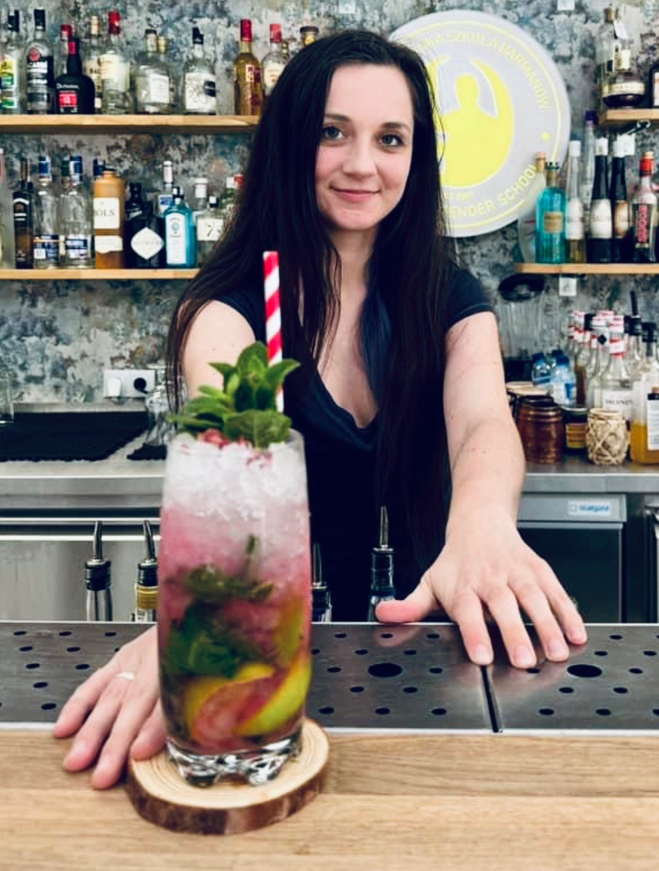 Uśmiechnięta barmanka prezentuje kolorowego drinka z kruszonym lodem, miętą i czerwoną słomką na drewnianej podkładce, w tle bar z alkoholem i logo szkoły barmańskiej.