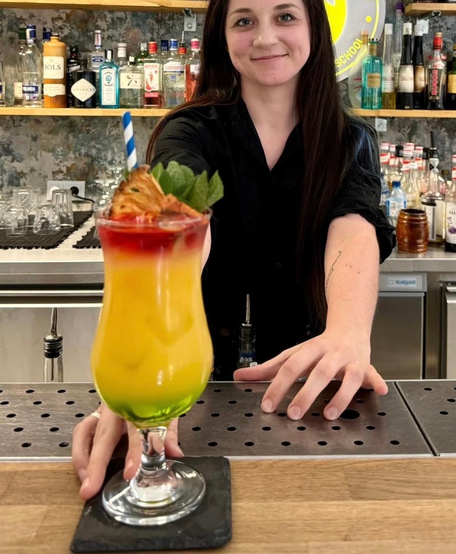 Uśmiechnięta barmanka serwuje kolorowy koktajl warstwowy z ananasem i miętą na barze, widoczne butelki z alkoholami w tle.