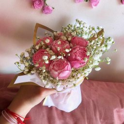 DIA MODA Patrycja Jelak - Dłoń trzyma delikatny bukiet z różowych róż i białej gipsówki, ozdobiony złotym papierem, na tle ściany z dekoracją z pojedynczych różowych kwiatów i różowego materiału.