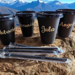 DIA MODA Patrycja Jelak - Cztery czarne kubki z imionami Wiktoria, Weronika, Jula i Angela napisanymi złotą czcionką, leżące na skale z kilkoma słomkami w foliowych opakowaniach, w tle górski krajobraz.