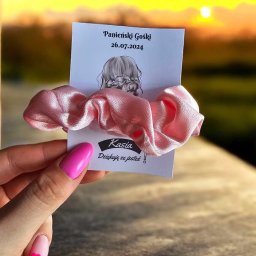 DIA MODA Patrycja Jelak - Różowa gumka do włosów z satyny, przymocowana do kartonika z napisem 'Panieński Gośki 26.07.2024' i rysunkiem fryzury, trzymana w dłoni z różowym manicure na tle zachodzącego słońca.