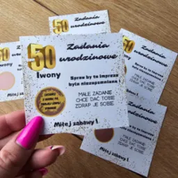 Karty ze zdrapkami z zadaniami urodzinowymi na 50 urodziny Iwony, trzymane w dłoni z różowym manicure na drewnianym tle.