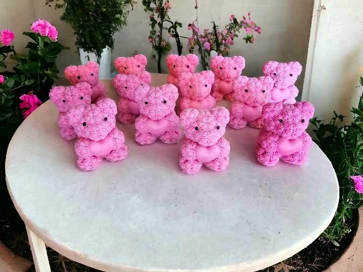 Rząd różowych misiaków z różyczek, każdy z serduszkiem, ustawionych na okrągłym stoliku, z kwiatami w tle.