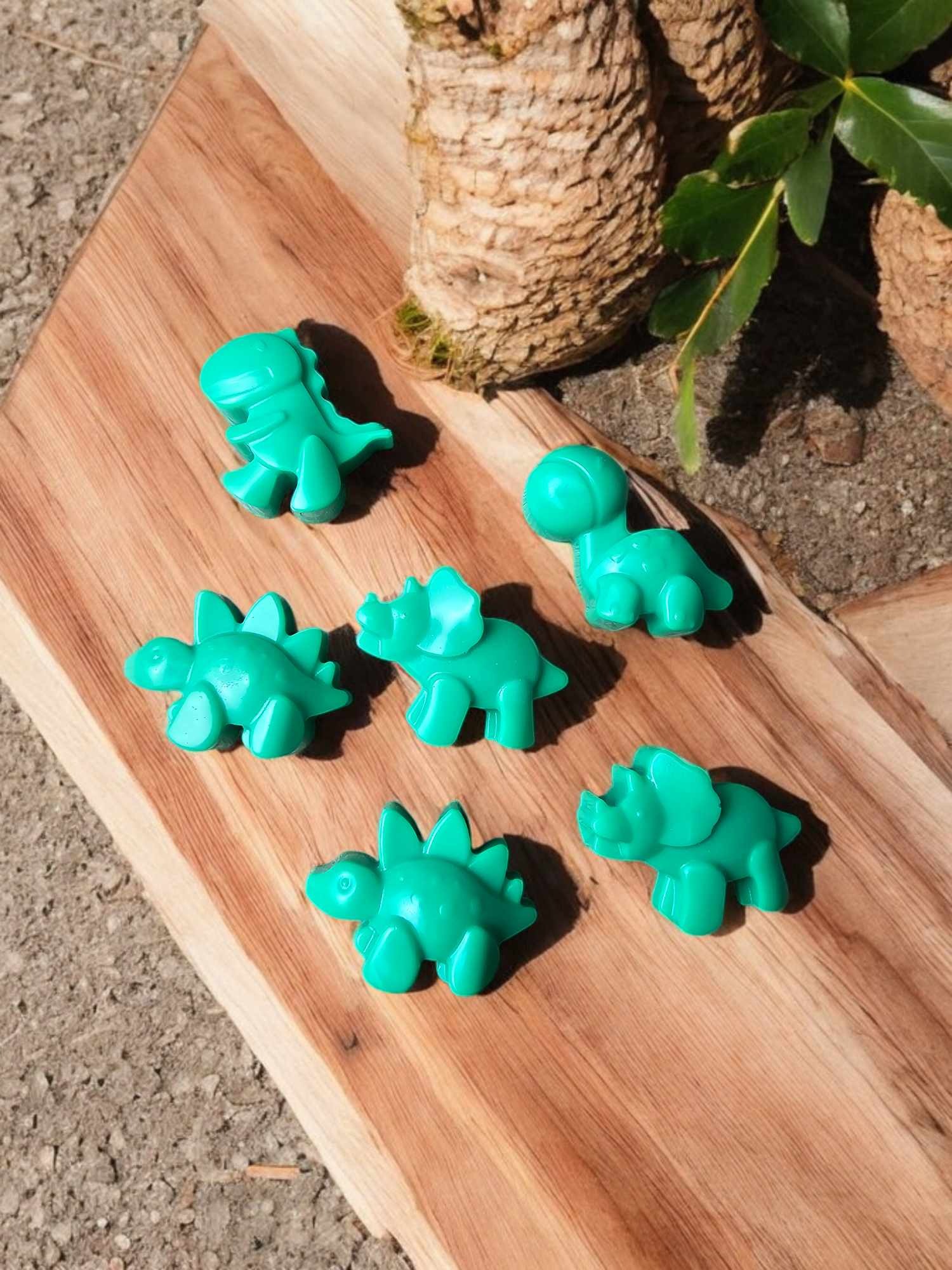 Sześć zielonych figurek dinozaurów z tworzywa sztucznego, różnych gatunków, ułożonych na drewnianej desce obok fragmentu kory drzewa i zielonych liści.