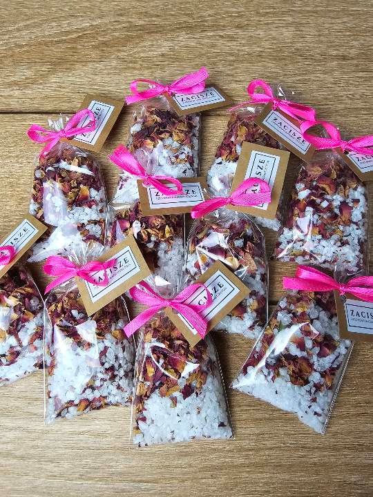 Seria przezroczystych woreczków z solą do kąpieli i suszonymi płatkami róż, przewiązanych różowymi wstążkami i oznaczonych etykietami na drewnianym stole.