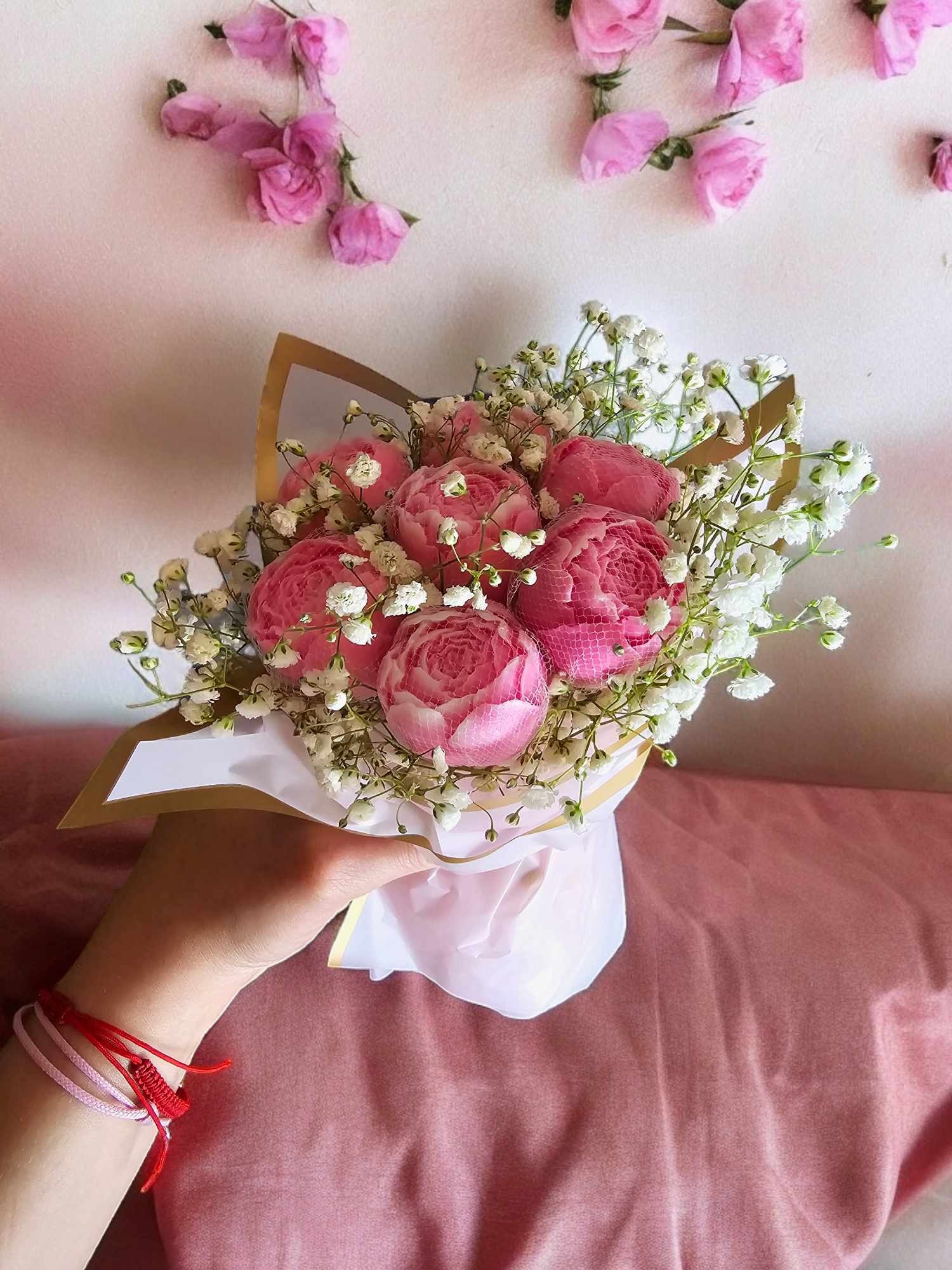 Dłoń trzyma delikatny bukiet z różowych róż i białej gipsówki, ozdobiony złotym papierem, na tle ściany z dekoracją z pojedynczych różowych kwiatów i różowego materiału.