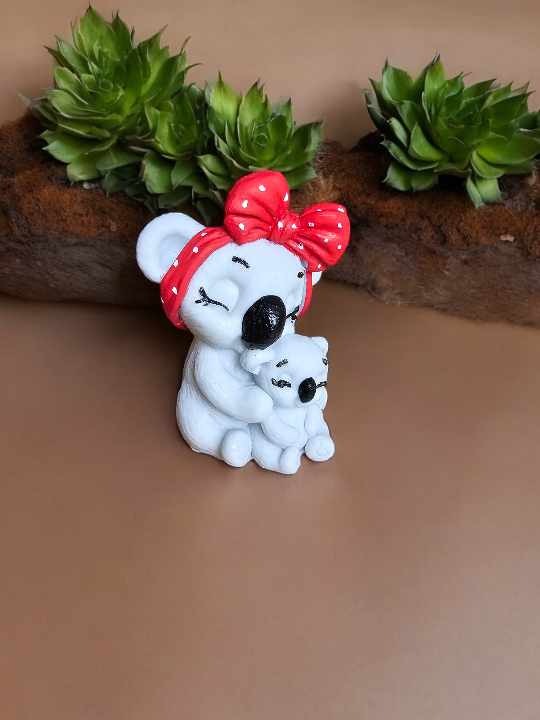 Ręcznie robiona figurka matki koali z dzieckiem, z czerwoną kokardą w białe kropki, umieszczona obok sukulentów na brązowym tle.