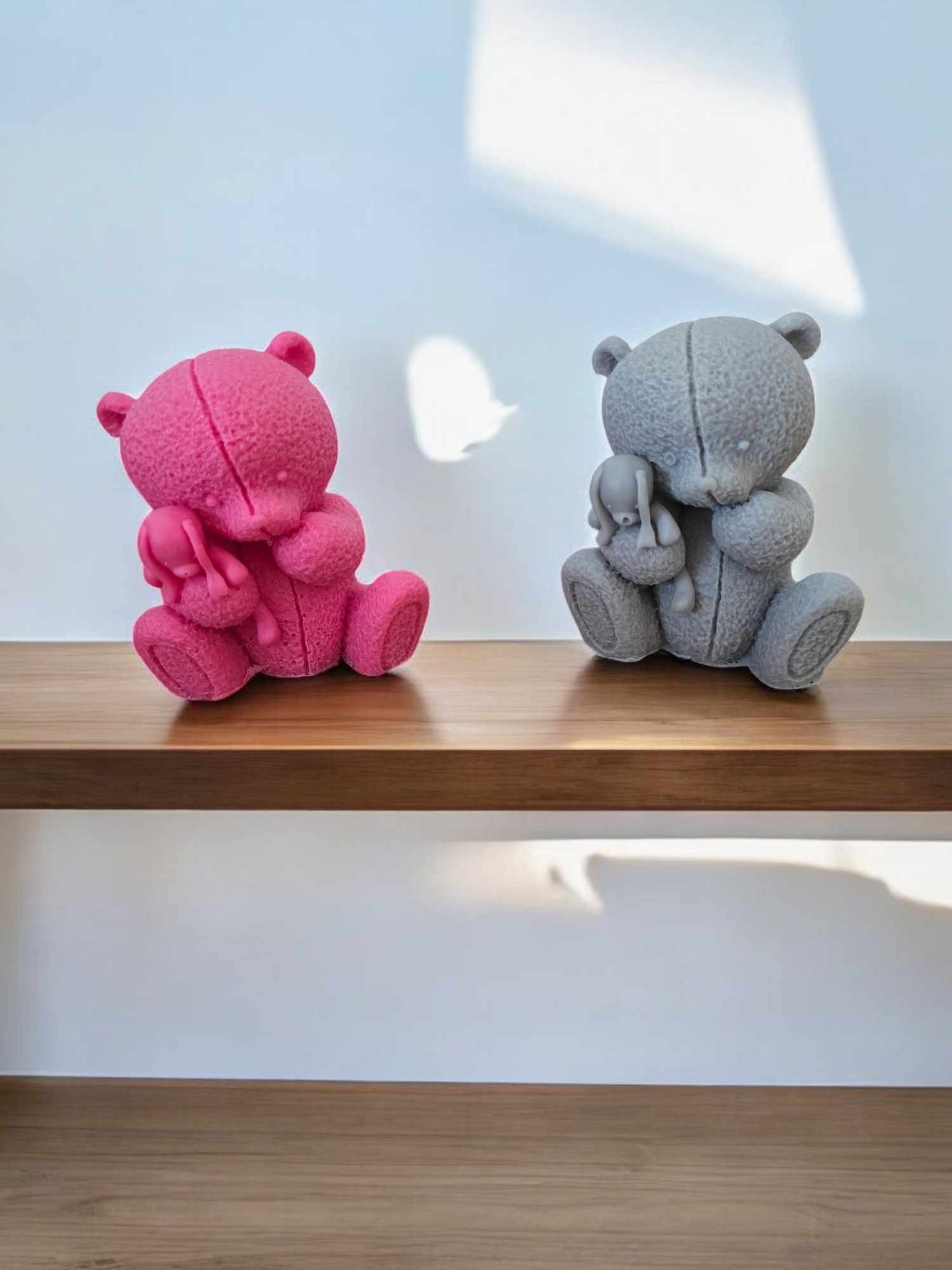 Dwa pluszowe misie, różowy i szary, siedzą na drewnianej półce pod białym tłem, każdy trzyma małą pluszową zabawkę, na ścianie widoczne cienie.
