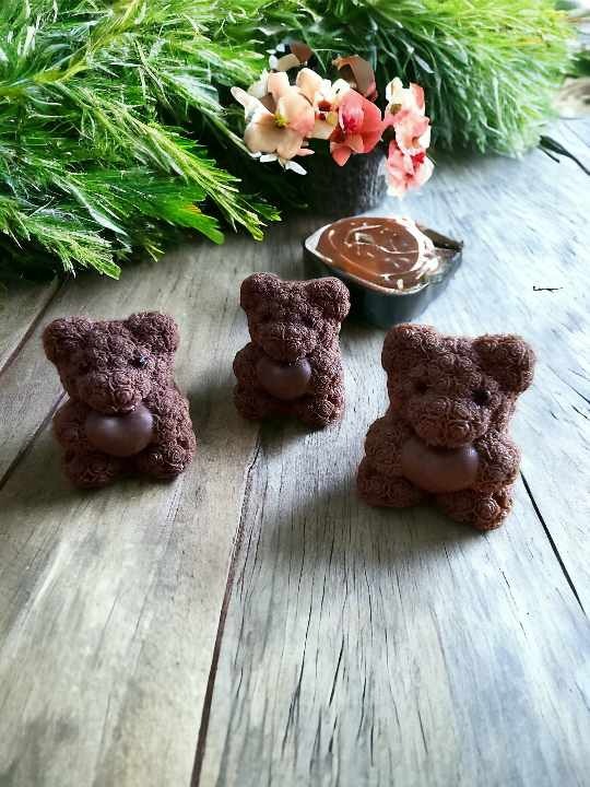 Trzy czekoladowe misie z sercami na drewnianym stole, w tle kompozycja kwiatowa i deser czekoladowy.