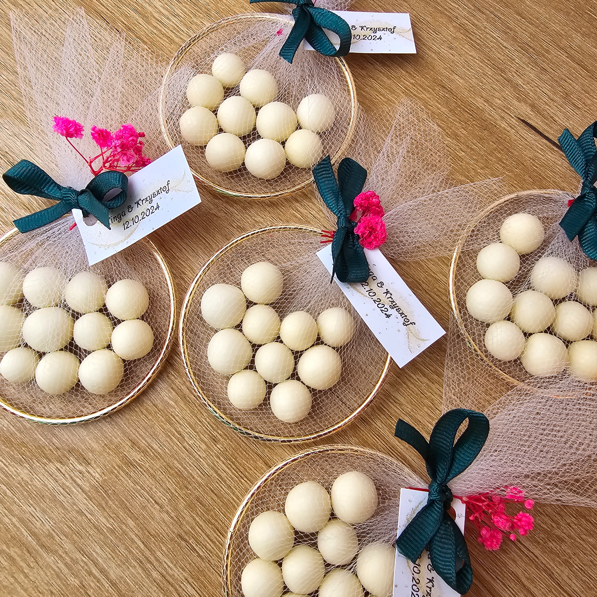 Dekoracyjne, złote koszyczki wypełnione białymi czekoladkami, owinięte tiulem i ozdobione zielonymi wstążkami i różowymi kwiatkami, z dołączonymi kartkami z imionami i datą ślubu, ułożone...