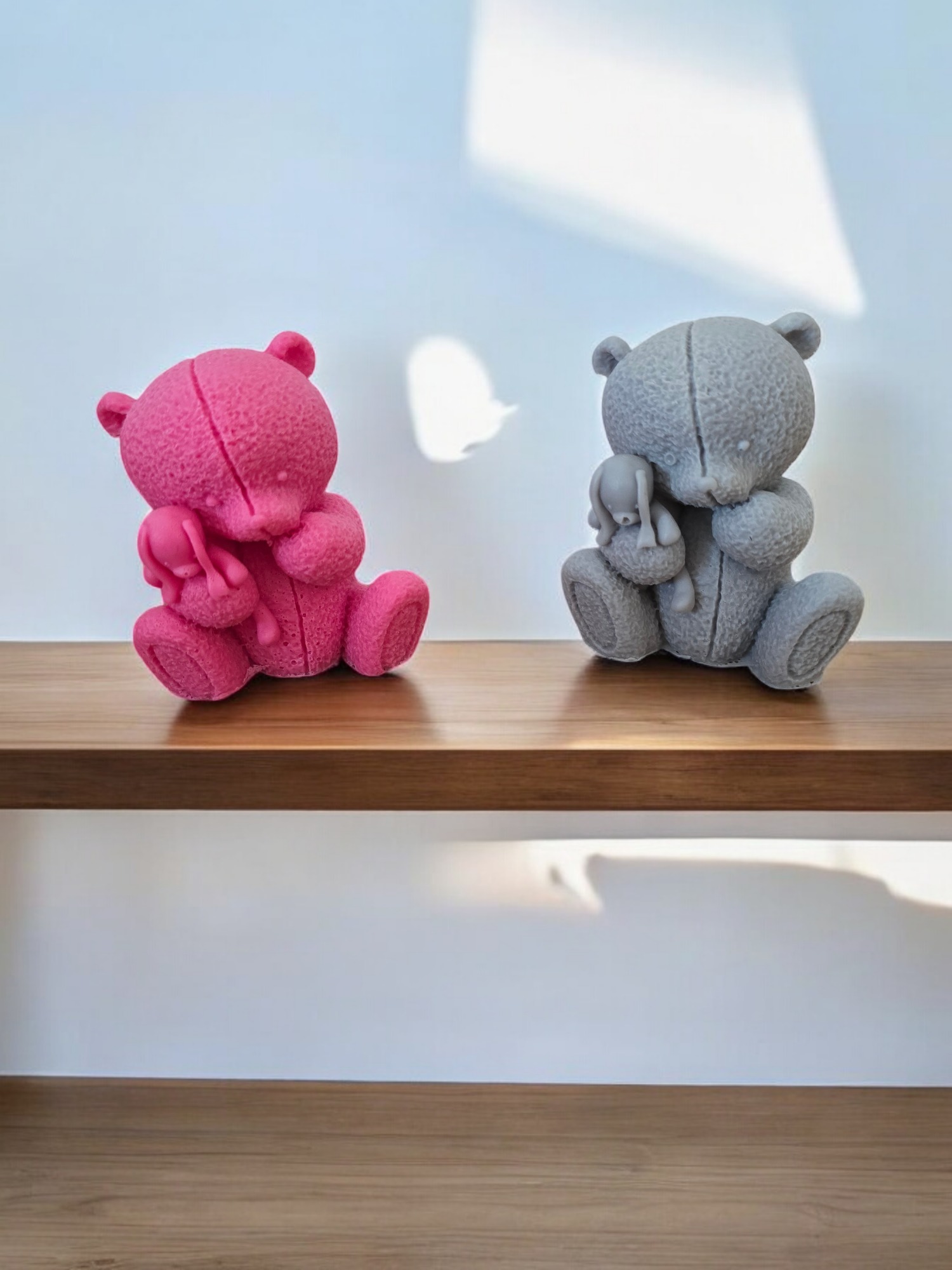 Dwa pluszowe misie na drewnianej półce: różowy trzyma króliczka, szary również trzyma króliczka, rzucając cień na ścianę.