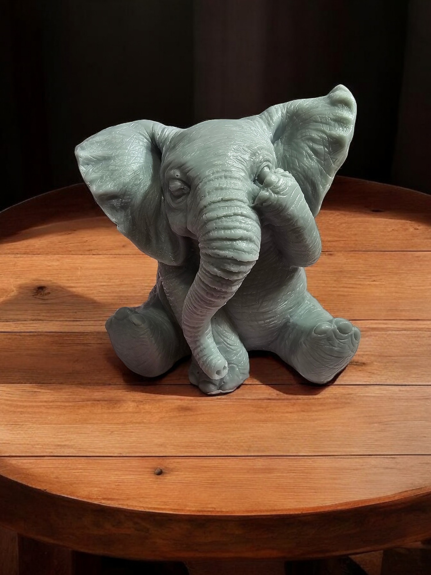 Szara, ręcznie wykonana figurka słonia z podpartą trąbą, siedząca na okrągłym, drewnianym stole z widocznymi słojami.
