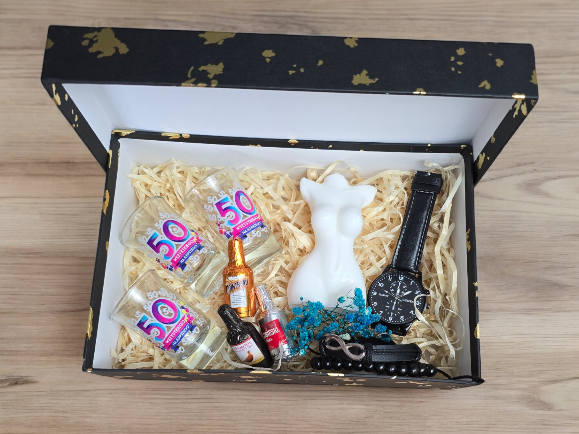 Otwarty czarny kosz prezentowy z złotymi zdobieniami, wypełniony słomkowym siankiem, zawierający trzy kieliszki z nadrukiem '50', miniaturowe buteleczki alkoholu, białą świecę w kształcie torsu...