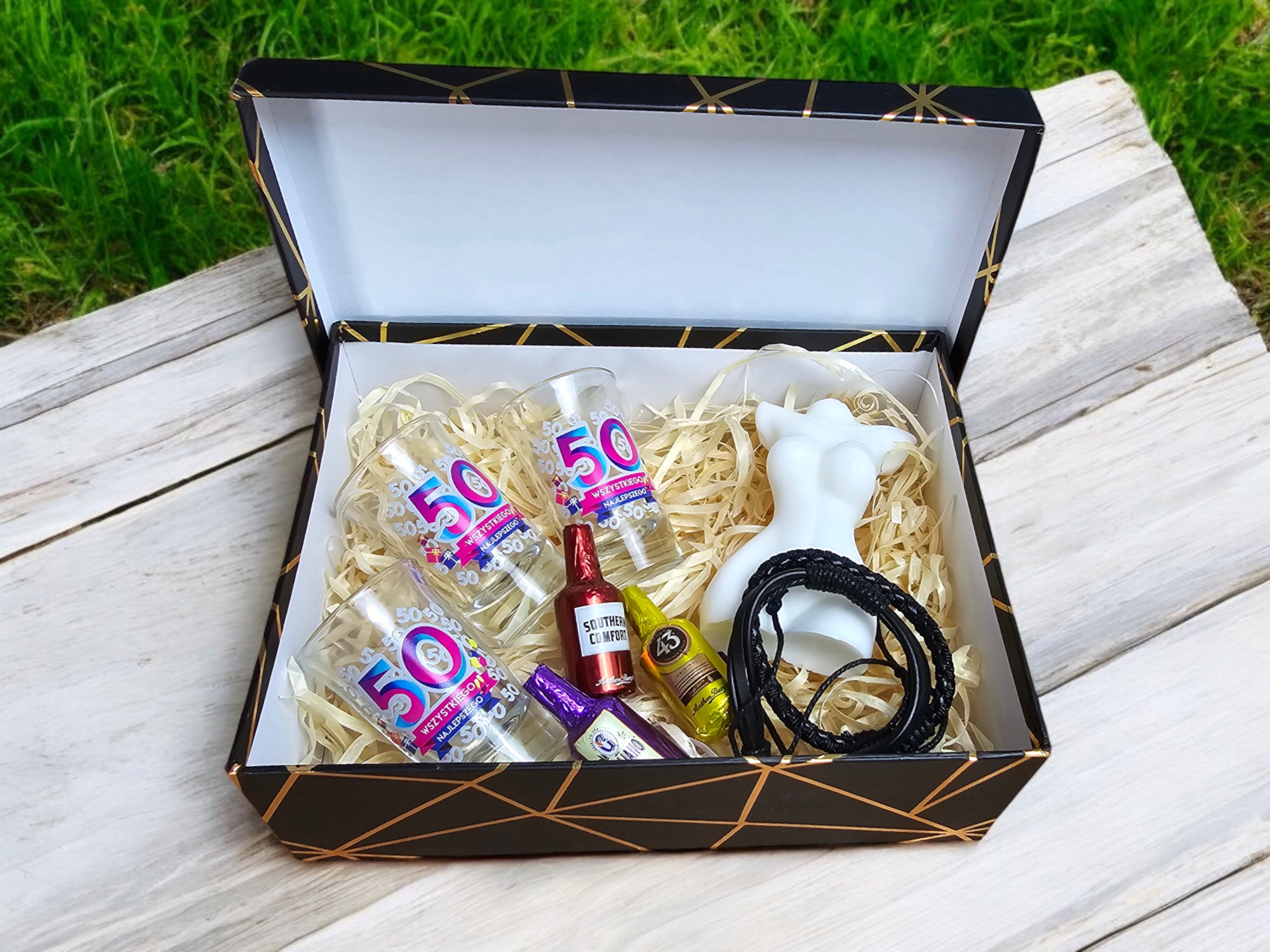 Otwarty czarny kosz prezentowy z geometrycznym wzorem, wypełniony czterema kieliszkami z nadrukiem '50 Wszystkiego Najlepszego', trzema czekoladowymi buteleczkami alkoholu, białą figurką kobiecego...