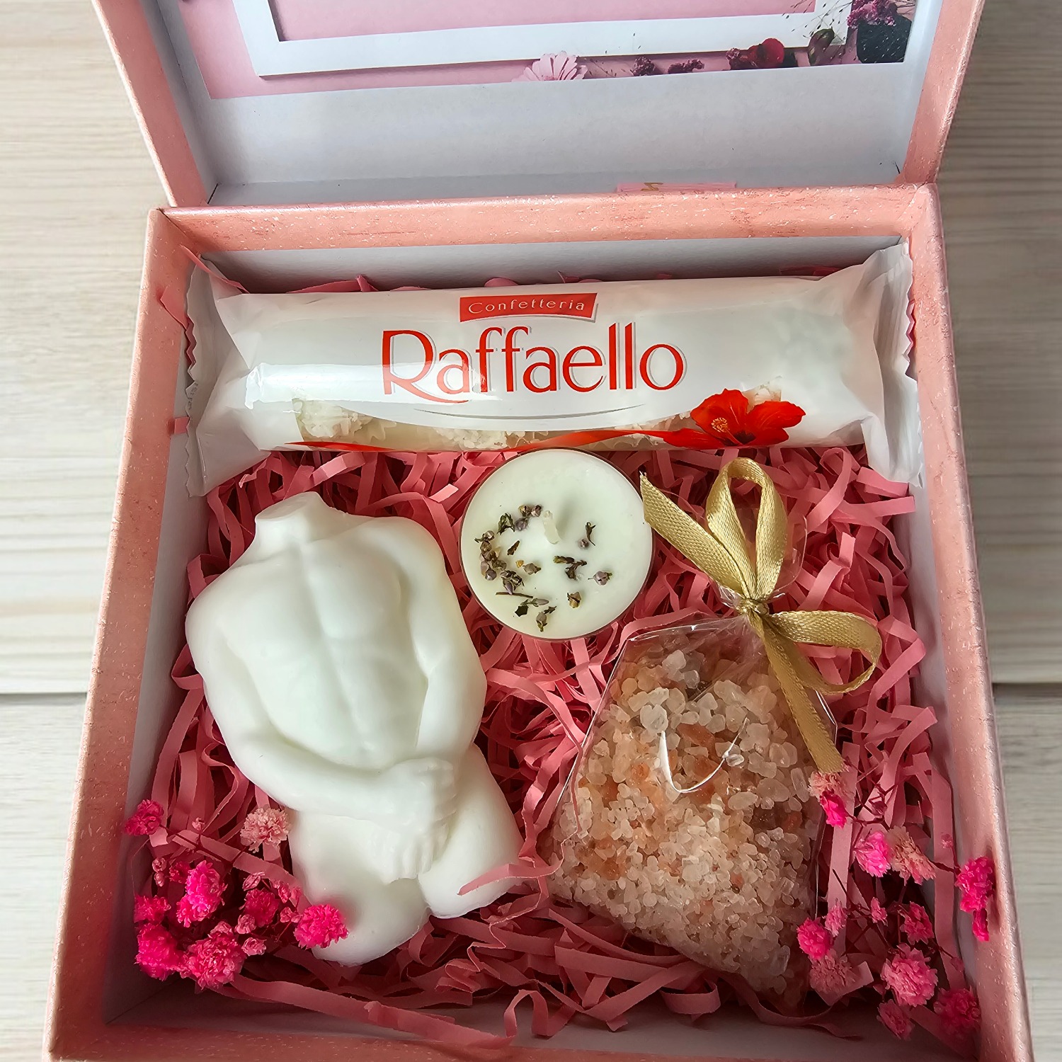 Otwarty, różowy kartonik z prezentami: batonik Raffaello, biała świeca w kształcie torsu, okrągła świeczka z ziołami i woreczek z solą do kąpieli, wszystko ułożone na różowej bibułce i ozdobione...