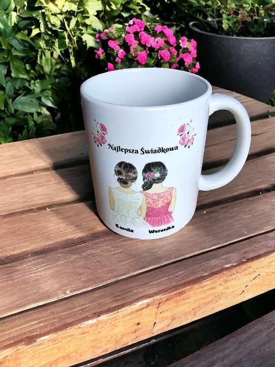 Biały kubek z nadrukiem 'Najlepsza Świadkowa' oraz ilustracją dwóch kobiet w sukienkach, stojący na drewnianym stole na tle zieleni i różowych kwiatów.