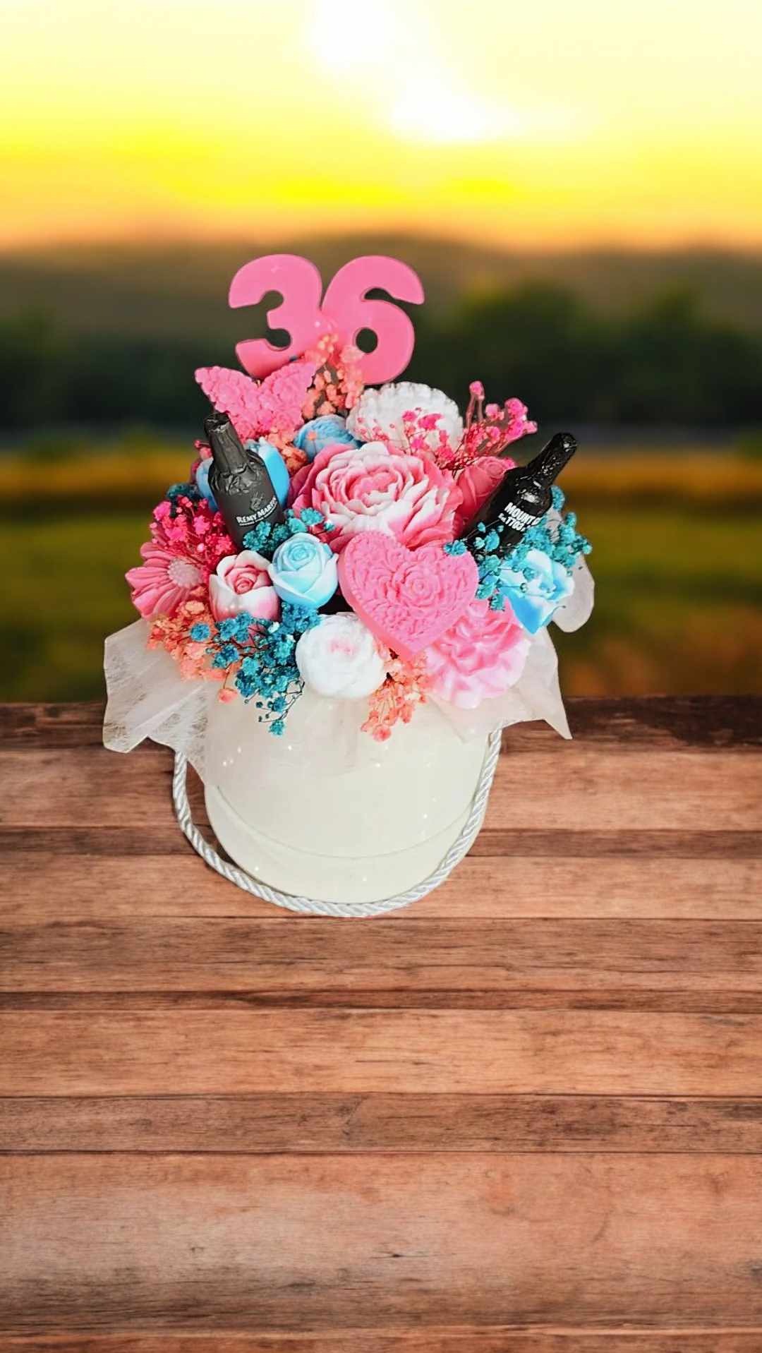 Ręcznie robiony kosz prezentowy z mydlanymi różami, sercami, miniaturowymi butelkami alkoholu i liczbą 36, umieszczony w białym pudełku z uchwytem sznurkowym, na drewnianym stole i rozmytym tle...