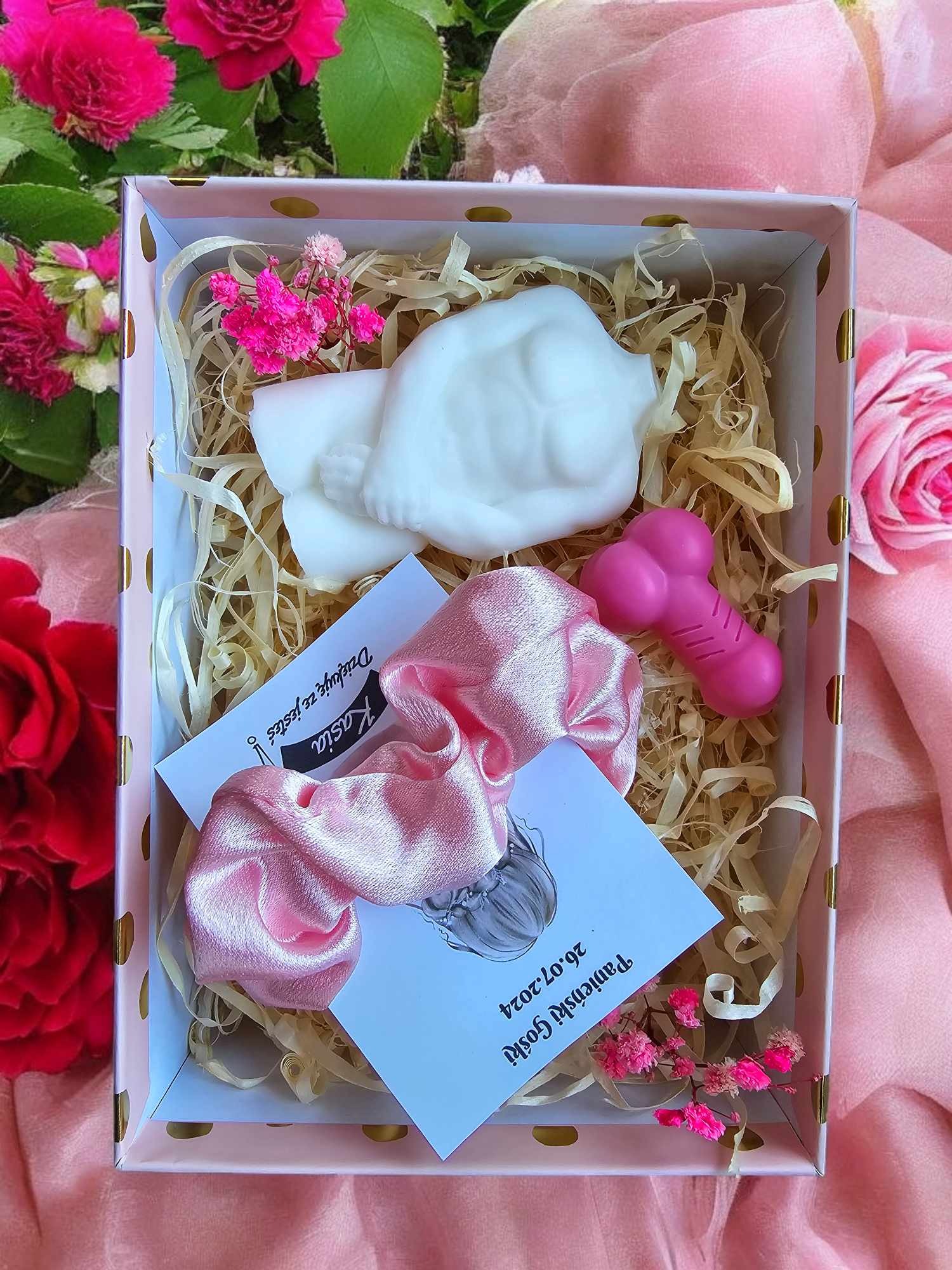 Otwarty ozdobny kartonik z zawartością: biała figurka dziecka z mydła, różowa gumka do włosów, różowy gadżet erotyczny, kartka z napisem 'Dziękuję że jesteś Kasia' i datą 26.07.2024, całość...