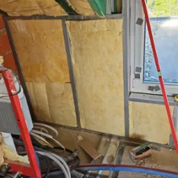 Montaż izolacji z wełny mineralnej na poddaszu, widoczne metalowe profile, drewniane belki stropowe pomalowane na zielono, poziomica, okno dachowe i instalacje sanitarne na podłodze.