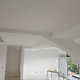 Sebastian Buda firma wykonczeniowa - Wnętrze w trakcie wykańczania, białe ściany i sufit z nietypową konstrukcją, widoczne przygotowane materiały na stole, duże okna z widokiem na zieleń.