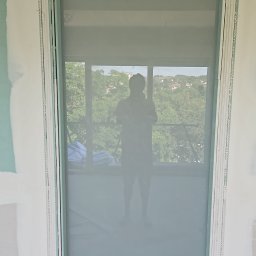 Sebastian Buda firma wykonczeniowa - Montaż płyty gipsowo-kartonowej wokół okna, widoczne profile i łączenia, w tle zarys sylwetki osoby i widok na zieleń.