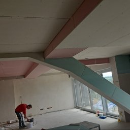 Sebastian Buda firma wykonczeniowa - Montaż konstrukcji z płyt gipsowo-kartonowych na suficie i ścianach, widoczny pracownik w czerwonej bluzie przygotowujący farbę, surowe wnętrze z oknami.