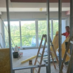 Sebastian Buda firma wykonczeniowa - Montaż ścianki działowej z płyty karton gips i ocieplenia z wełny mineralnej, widoczna konstrukcja metalowa, drabina i pracownik w czerwonej koszulce na tle okna z widokiem na zieleń.