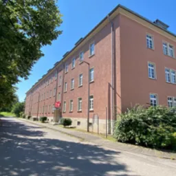 Trzypiętrowy budynek z elewacją w kolorze terakoty, widoczny z poziomu ulicy w słoneczny dzień, z zielenią drzew i krzewów w tle.