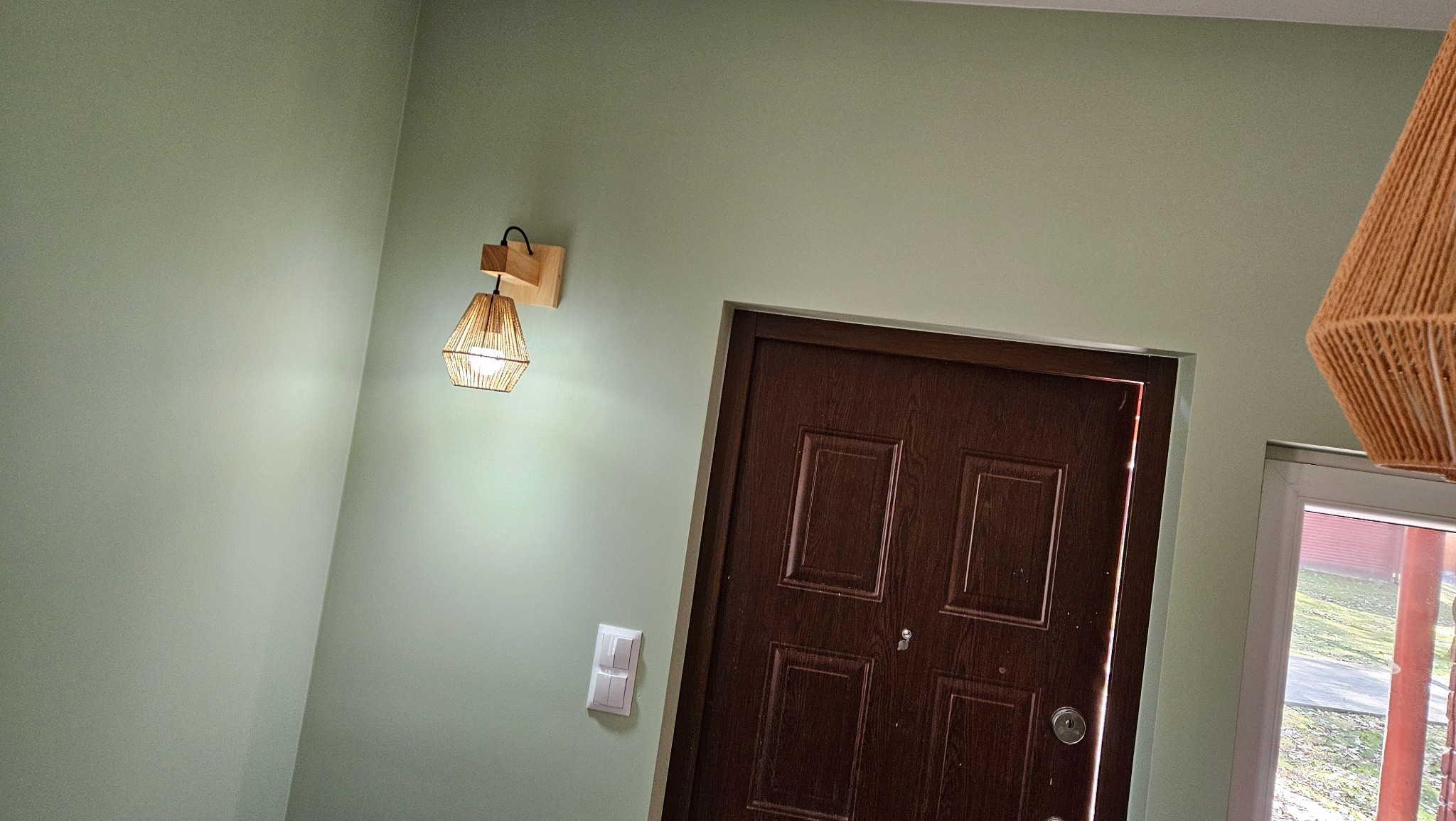 Ściana pomalowana na pistacjowy kolor z brązowymi drzwiami, drewniany kinkiet ze sznurkowym abażurem i widoczny fragment wiszącej lampy w rogu kadru.