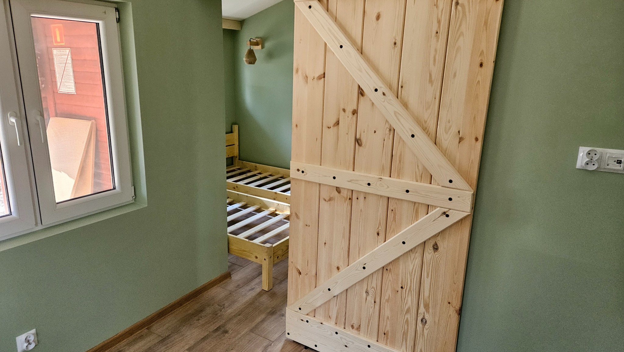 Drewniane przesuwne drzwi stodołowe, częściowo otwarte, odsłaniające wnętrze pokoju z drewnianym łóżkiem piętrowym na tle zielonej ściany.