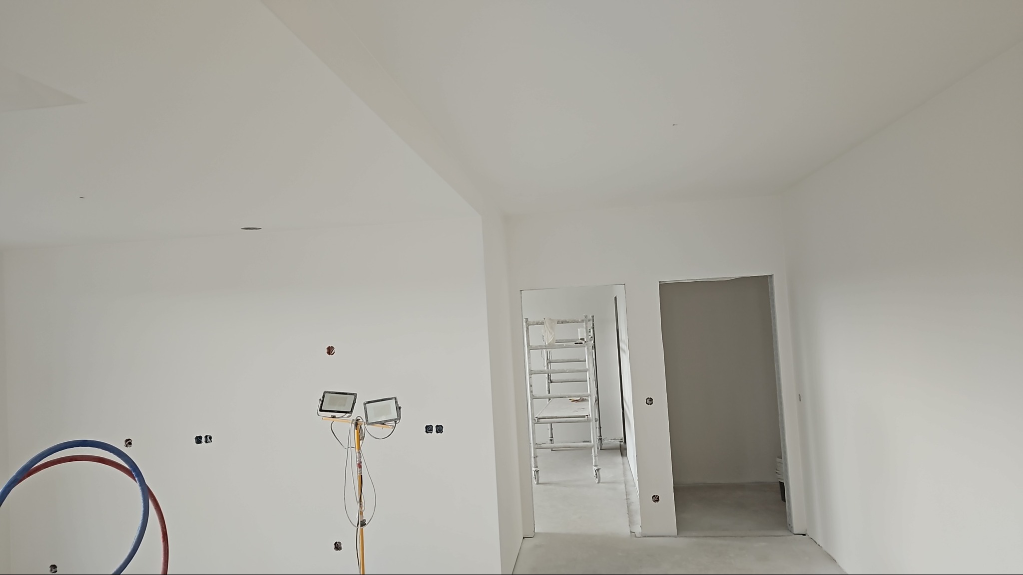 Wnętrze w trakcie malowania na biało, widoczne ściany po gładziowaniu, instalacja elektryczna, rury instalacji wodnej oraz drabina malarska w przejściu do innego pomieszczenia.