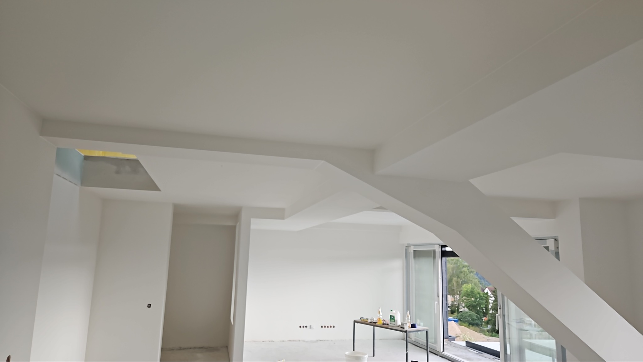 Wnętrze w trakcie wykańczania, białe ściany i sufit z nietypową konstrukcją, widoczne przygotowane materiały na stole, duże okna z widokiem na zieleń.