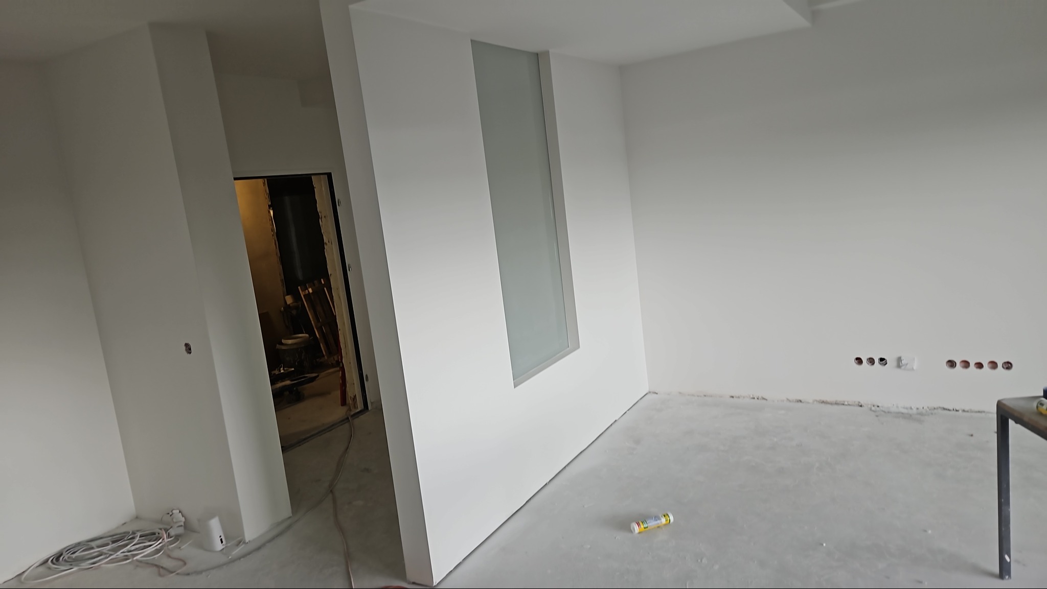 Wnętrze w trakcie wykańczania, z widocznymi przewodami elektrycznymi, puszkami pod gniazdka i listwą przypodłogową. Na podłodze leży tuba z klejem. Ściany są białe, z wstawionym pionowym, matowym...