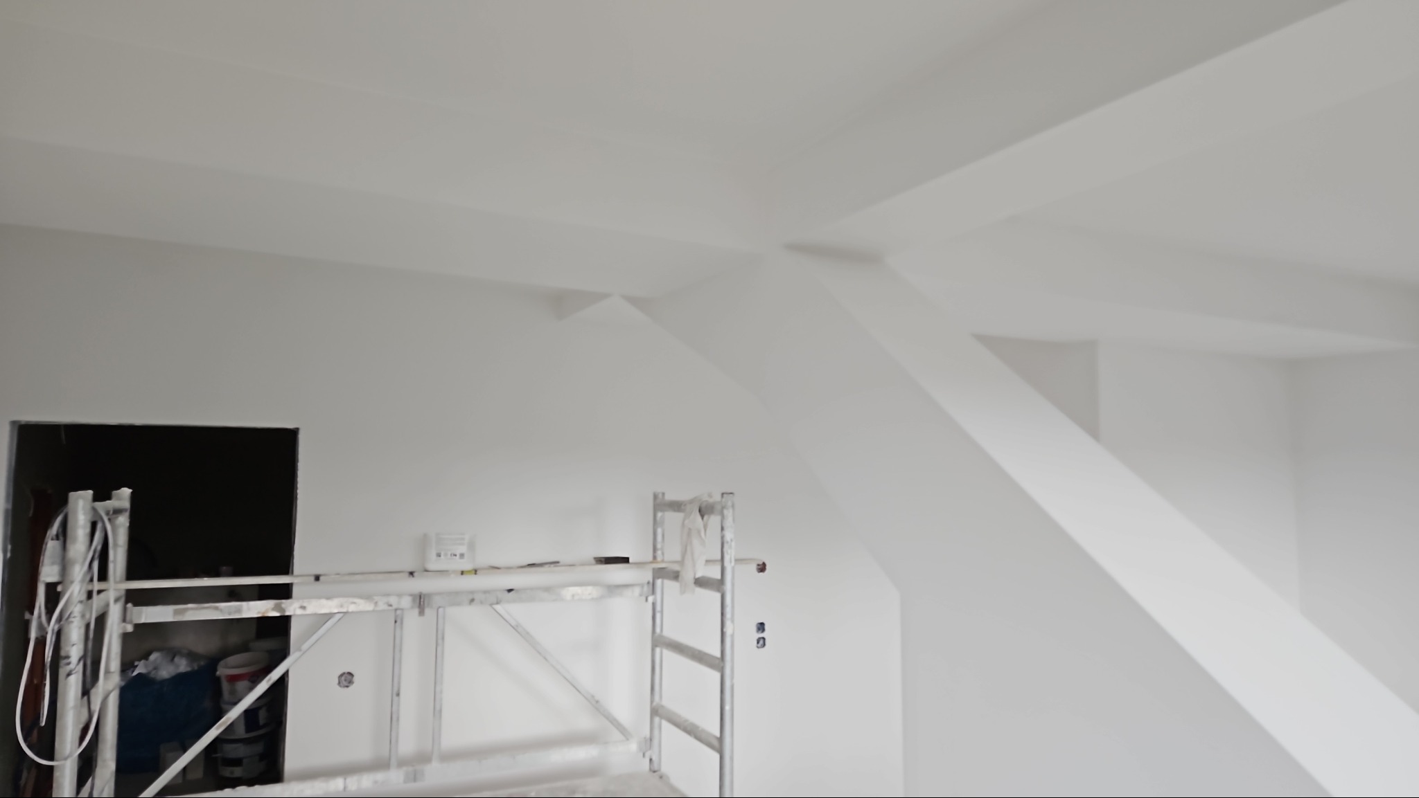 Świeżo pomalowane na biało poddasze ze skosami i belkami, widoczne rusztowanie malarskie i otwór drzwiowy.