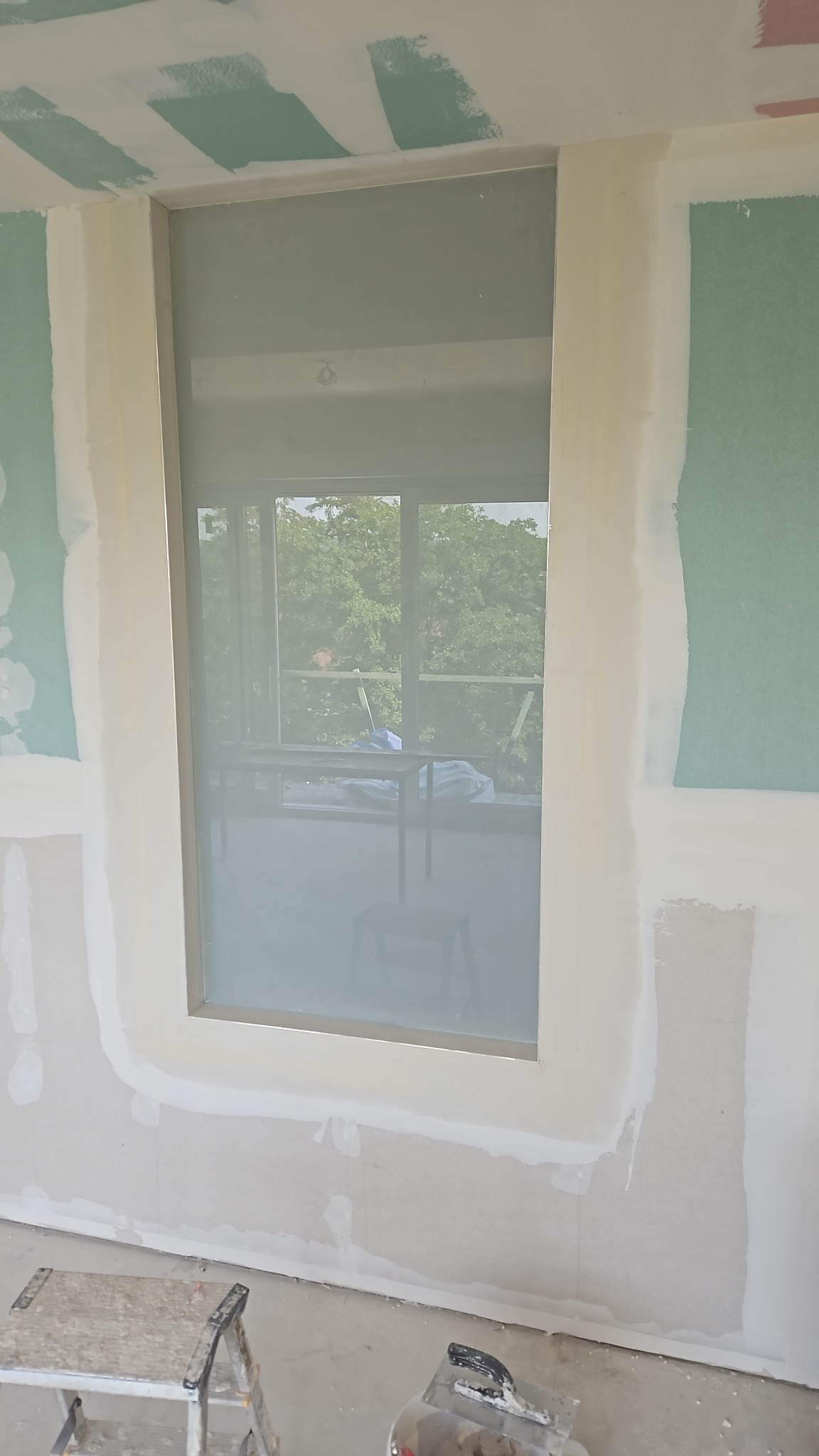 Ściana z oknem w trakcie szpachlowania, widoczne łączenia płyt gipsowych, szpachelka i drabina w dolnej części kadru.