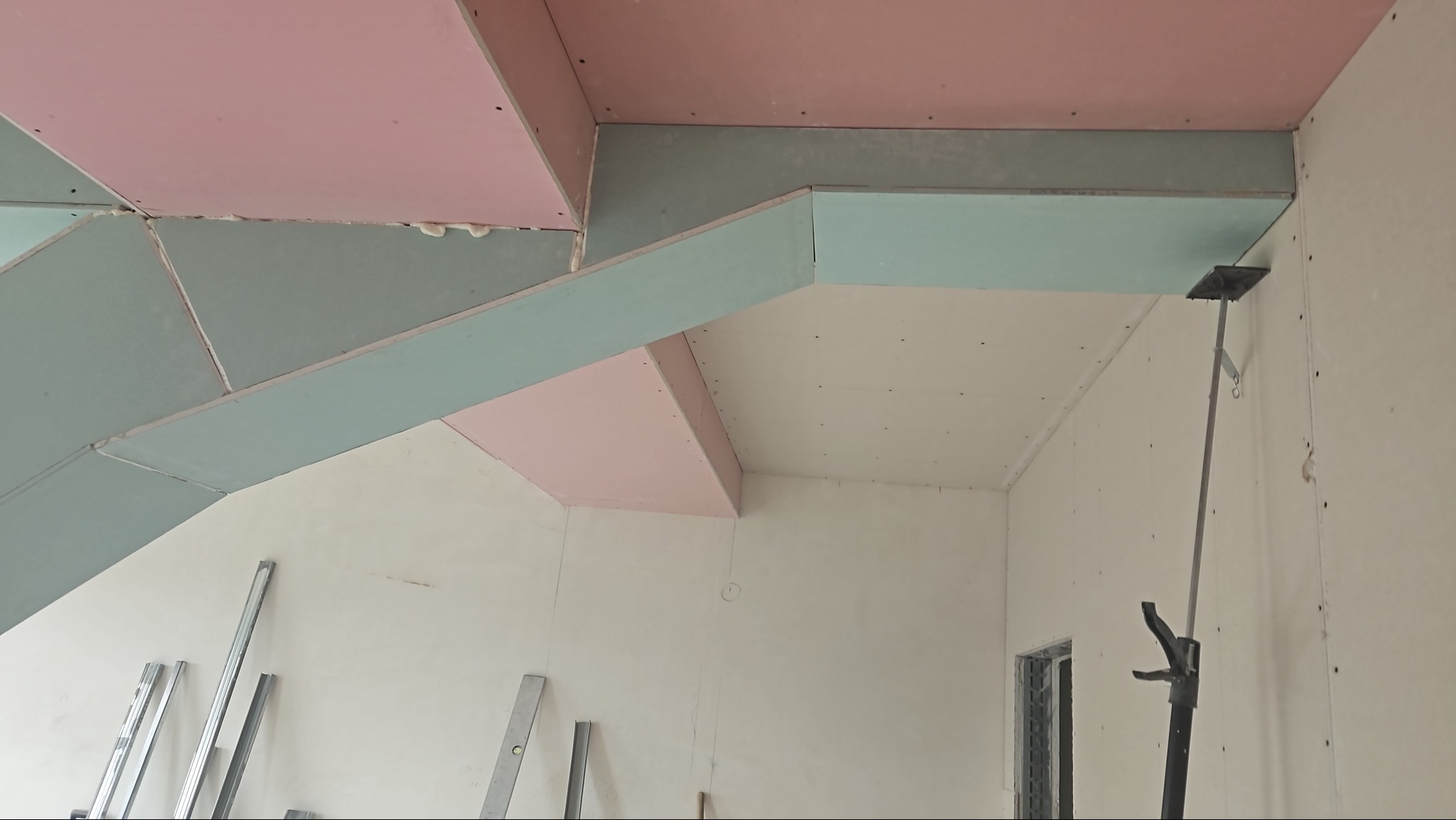 Montaż sufitu podwieszanego z płyt w odcieniach różu i szarości, widoczne profile i narzędzia, w tle przygotowane ściany do dalszych prac wykończeniowych.