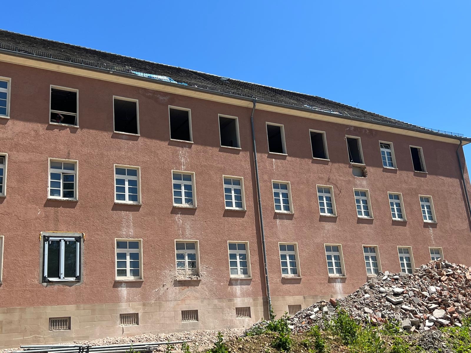 Trzypiętrowy budynek z różową elewacją w trakcie remontu, widoczne okna bez szyb, gruzowisko u podnóża ściany, błękitne niebo w tle.