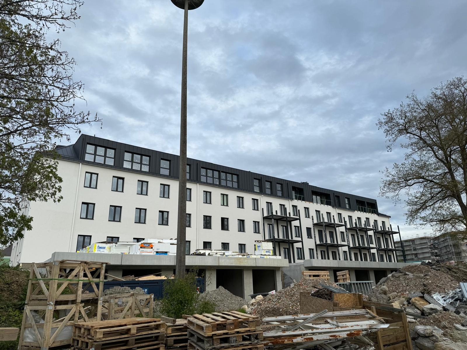 Wielopiętrowy budynek w trakcie wykańczania, z widocznymi materiałami izolacyjnymi na balkonie i stertą gruzu oraz palet przed budynkiem, pochmurne niebo w tle.