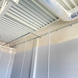 Fifi-Spaw - Kable w korytkach i rury pod sufitem w nowym budynku. Szare ściany w połowie pomalowane. Widok na sufit z blachy trapezowej i konstrukcję stalową.