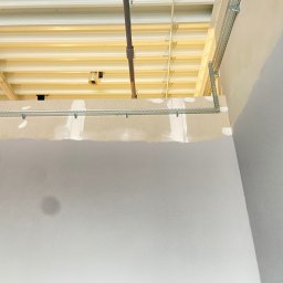 Fifi-Spaw - Szara ściana z metalowymi elementami mocującymi pod sufitem. Widoczne rury i kable pod stropem. Prace wykończeniowe wewnątrz budynku.