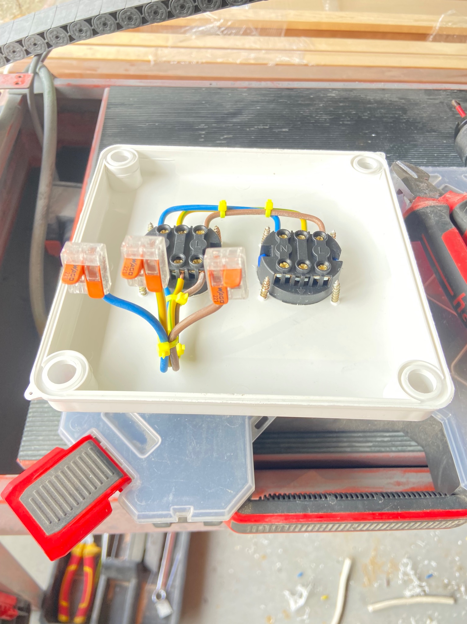 Wnętrze białej puszki elektrycznej z podłączonym gniazdem przy użyciu złączek WAGO i przewodów w kolorach: niebieskim, żółto-zielonym i brązowym, zabezpieczonych żółtymi opaskami zaciskowymi.