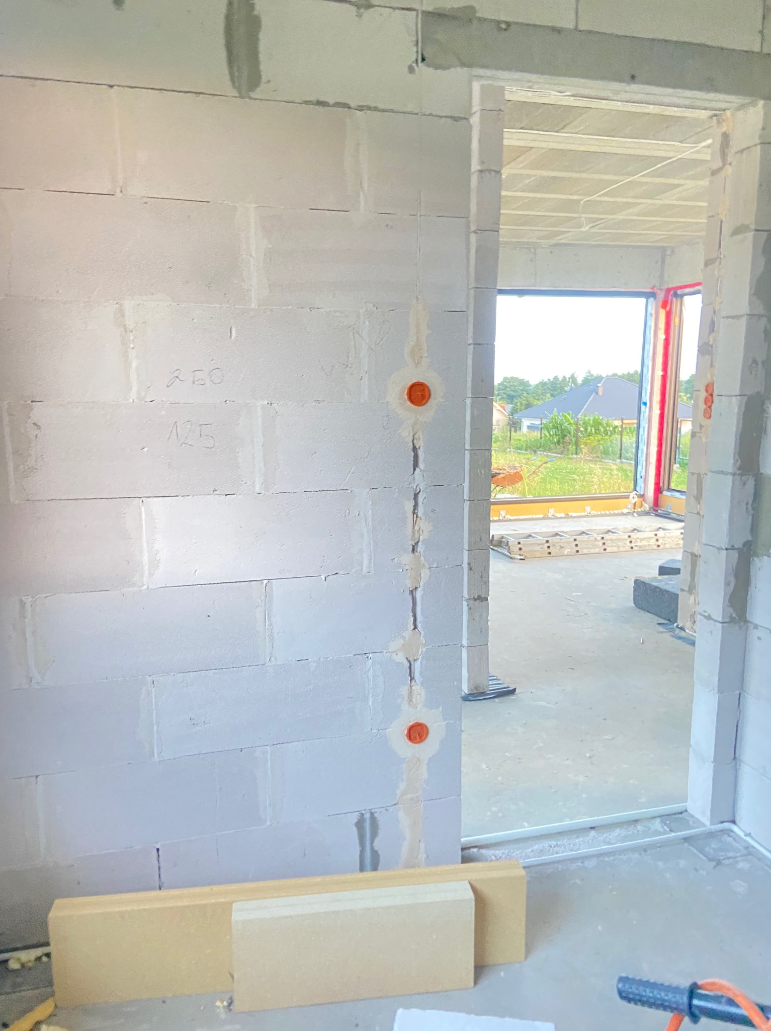 Ściana z betonu komórkowego w trakcie budowy, z widocznymi puszkami instalacyjnymi w pionowym wyżłobieniu, przygotowanymi do rozprowadzenia przewodów. Na ścianie widoczne odręczne notatki. W tle...