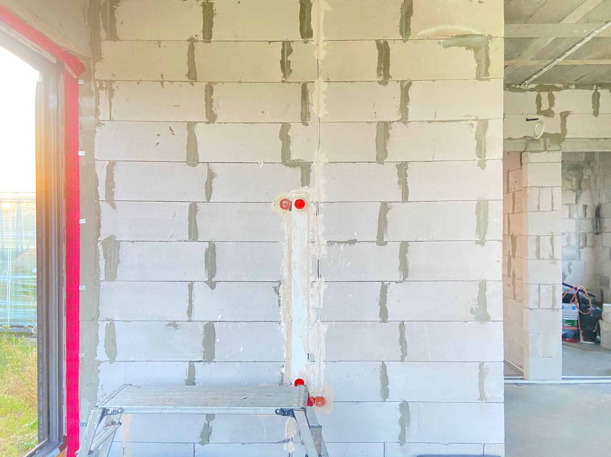 Ściana z betonu komórkowego w trakcie budowy, z wyżłobieniami na przewody elektryczne i zamontowanymi puszkami instalacyjnymi, widoczne rusztowanie aluminiowe i fragment okna.