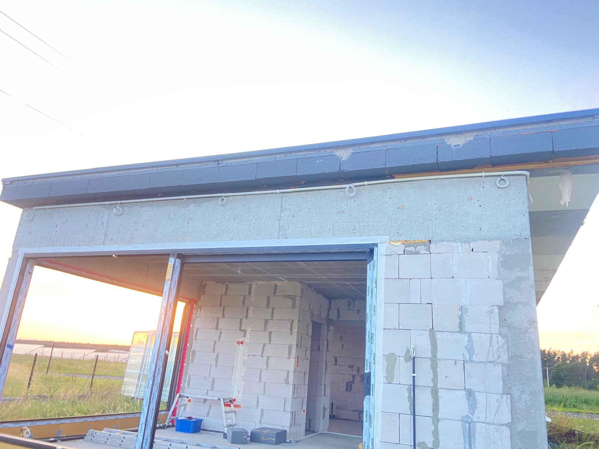 Surowy budynek w trakcie budowy z widocznymi przewodami elektrycznymi zamocowanymi na zewnątrz, z oknami z profilami aluminiowymi, w tle zachód słońca.