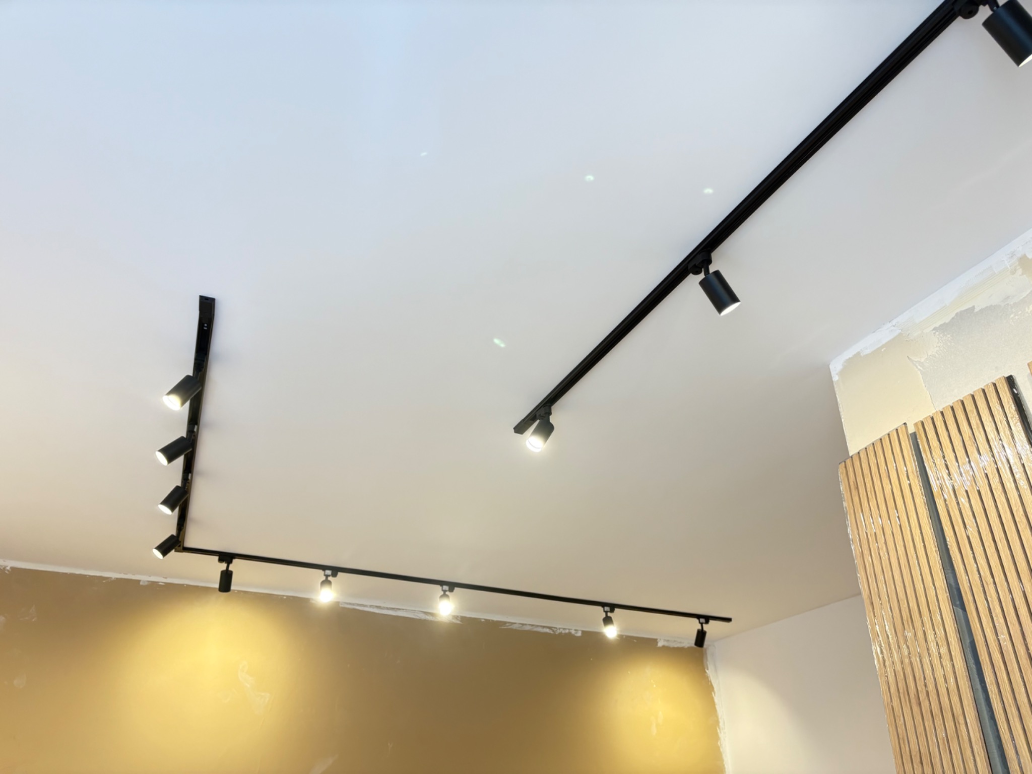 Nowoczesny system szynowy z czarnymi reflektorami sufitowymi, oświetlający wnętrze w trakcie wykończenia. Widoczne płyty dekoracyjne na ścianie.