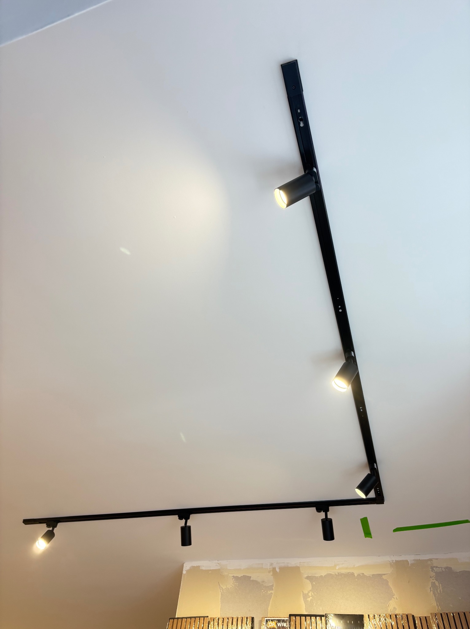 Czarna, szynowa instalacja oświetleniowa z trzema reflektorami na białym suficie, z widocznymi śladami prac wykończeniowych na ścianie.